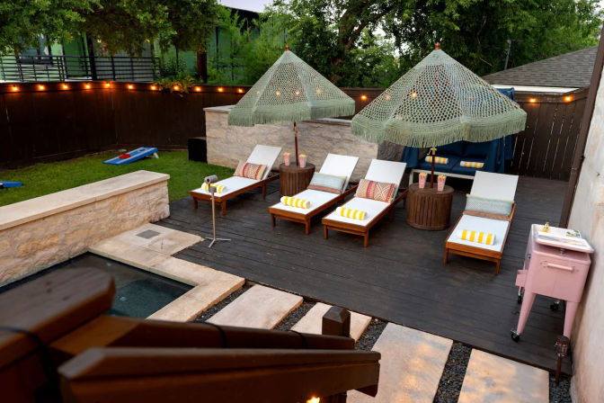 Backyard patio with a small plunge pool and stone edging, four wooden sun loungers with yellow-striped towels under green fringed umbrellas on a dark wooden deck, string lights along the fence, cornhole boards on the lawn and a pink rolling cooler.