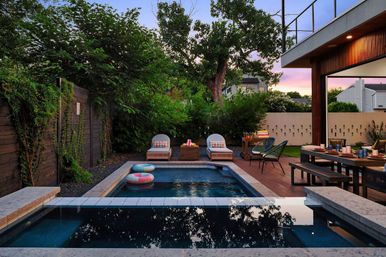 Modern backyard pool oasis at sunset — rectangular swimming pool with raised spa and colorful floats, wooden deck with lounge chairs and an outdoor dining table surrounded by lush privacy greenery.