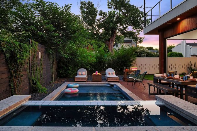 Modern backyard pool oasis at sunset — rectangular swimming pool with raised spa and colorful floats, wooden deck with lounge chairs and an outdoor dining table surrounded by lush privacy greenery.