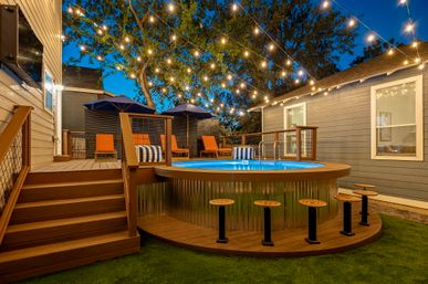 Cozy residential backyard patio at dusk with a round above‑ground pool on a wooden deck, bar stools at the pool edge, orange lounge chairs under navy umbrellas and warm string lights overhead.