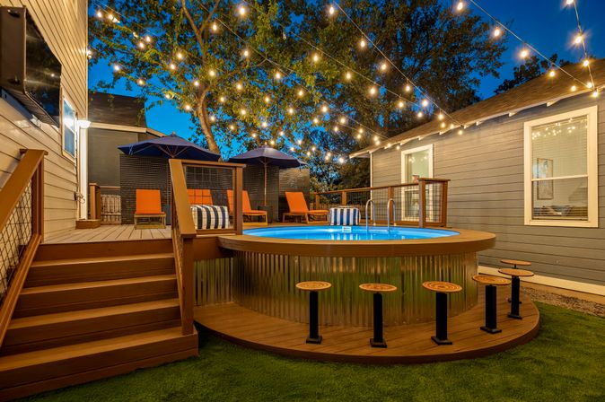 Cozy residential backyard patio at dusk with a round above‑ground pool on a wooden deck, bar stools at the pool edge, orange lounge chairs under navy umbrellas and warm string lights overhead.