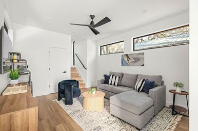 Bright modern living room with gray L-shaped sectional, patterned rug, round wooden coffee table, navy accent chairs, wood floors, narrow high windows and black ceiling fan