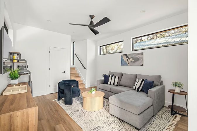 Bright modern living room with gray L-shaped sectional, patterned rug, round wooden coffee table, navy accent chairs, wood floors, narrow high windows and black ceiling fan