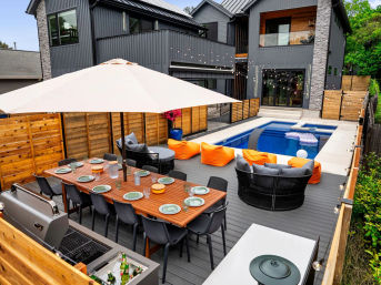 Modern backyard deck with a long outdoor dining table under a large umbrella, built-in grill, cozy lounge seating with bright orange cushions, string lights, and a rectangular pool beside a dark-gray two-story house.