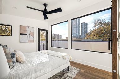 Bright modern downtown apartment bedroom with white bed and textured pillows, large windows framing city high-rises and fall foliage, wood floors, black ceiling fan and abstract wall art.