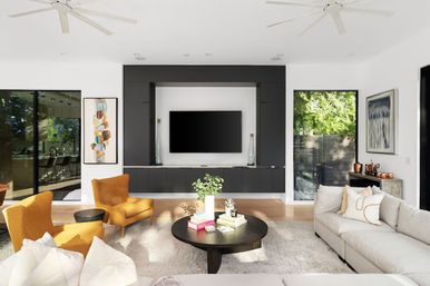 Sunlit modern living room with black built-in media wall and wall-mounted TV, light gray sectional, two mustard accent chairs, round coffee table with books and plant, large glass doors opening to a green backyard.