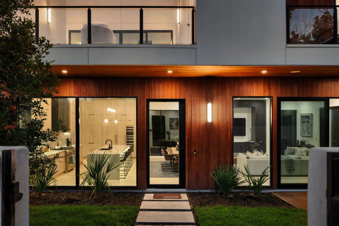 Contemporary two-story house at dusk — warm vertical wood-clad facade with floor-to-ceiling glass showing a lit open-plan kitchen and cozy living room, balcony above and stepping-stone front path.