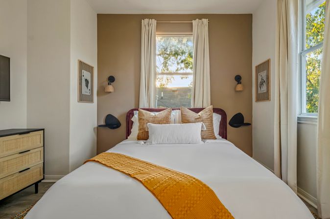 Sunlit cozy bedroom with white bedding, mustard throw, layered pillows, velvet headboard, rattan dresser and window view of trees