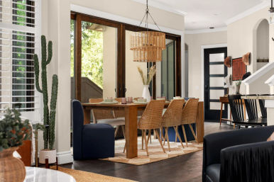 Sunlit modern open-concept dining room with wooden table, six rattan chairs and navy upholstered bench, woven pendant light, large sliding glass doors to patio, potted cactus and neutral décor.