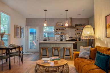 Cozy, sunlit open-plan living room and kitchen in a modern suburban home — warm rust velvet sofa with teal throw pillows, rattan round coffee table, wooden island with three bar stools, white subway tile backsplash, stainless steel range hood, pendant lights, dining nook by window, wood floors and glass door to a patio.