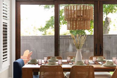 Sunlit dining room with wooden table set for six, woven rattan chairs, ceramic place settings and pink glass tumblers, tall white vase of dried grasses, layered wicker pendant light, and sliding glass doors opening to a leafy backyard.