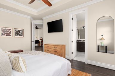 Bright contemporary bedroom interior with beige walls and dark hardwood floors, white bedding with striped pillows, wooden dresser beneath a wall-mounted flat-screen TV, arched full-length mirror reflecting a lamp and plant, open doors to an ensuite bathroom and home office, ceiling fan and framed wall art.