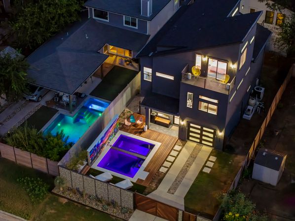 Aerial night view of a modern suburban three-story home with a glowing purple plunge pool on a wooden deck, balcony with yellow chairs, adjacent turquoise pool, lit driveway and landscaped backyard.