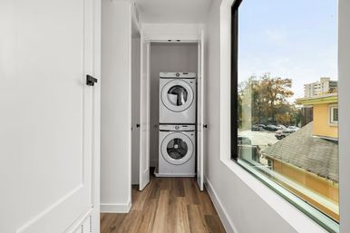 Bright modern apartment hallway with stacked front-load washer and dryer in an open closet, wood-look flooring and a large window overlooking nearby buildings and a parking lot.