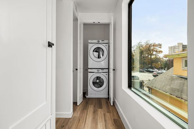 Bright modern apartment hallway with stacked front-load washer and dryer in an open closet, wood-look flooring and a large window overlooking nearby buildings and a parking lot.