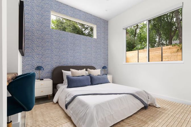 Modern bedroom with blue scallop-pattern accent wall, low bed with navy pillows and white nightstands, large window showing wooden fence and trees.