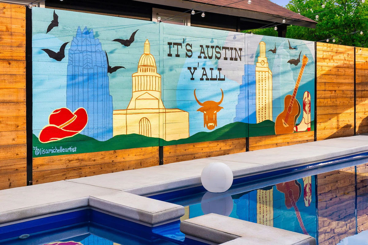 Vibrant mural on a wooden fence beside a pool reading 'It's Austin Y'all,' showing the Austin skyline with the Texas Capitol dome, skyscrapers, a longhorn skull, cowboy hat, guitar and flying bats reflected in the water.