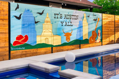 Vibrant mural on a wooden fence beside a pool reading 'It's Austin Y'all,' showing the Austin skyline with the Texas Capitol dome, skyscrapers, a longhorn skull, cowboy hat, guitar and flying bats reflected in the water.