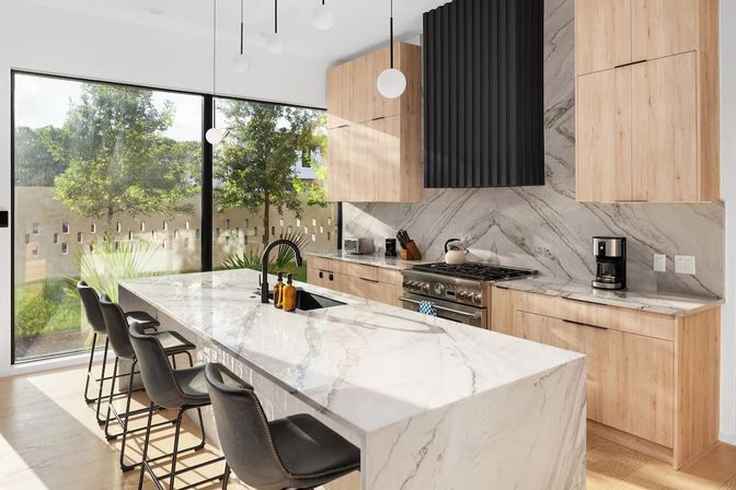 Bright modern kitchen with floor-to-ceiling windows and leafy yard views, white marble waterfall island and breakfast bar with black faucet and four leather stools, light wood cabinets, marble backsplash, stainless steel range and pendant globe lights.