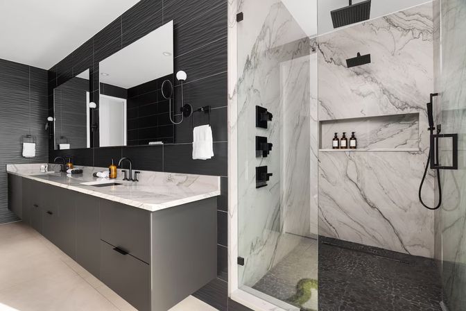 Modern spa-like bathroom with marble walk-in shower and glass enclosure, dark textured tile walls, double marble vanity, large mirrors and black matte fixtures.
