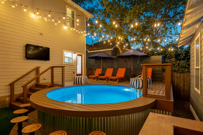 Cozy backyard deck at dusk with a round above-ground illuminated pool, hanging string lights, orange lounge chairs under umbrellas, striped poufs and wooden bar stools.