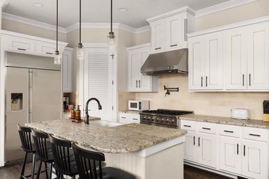 Bright modern white kitchen with granite island and breakfast bar, stainless steel fridge and professional range with hood, three black bar stools and pendant lights.