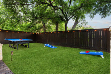 Fenced residential backyard with lush green turf, portable ping-pong table and two blue cornhole boards, warm string lights along a dark wooden fence and large shade trees overhead — casual outdoor games and entertaining space.
