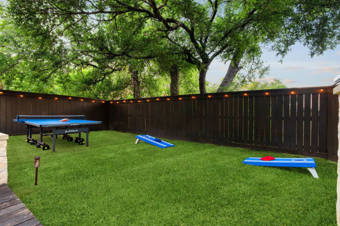 Fenced residential backyard with lush green turf, portable ping-pong table and two blue cornhole boards, warm string lights along a dark wooden fence and large shade trees overhead — casual outdoor games and entertaining space.