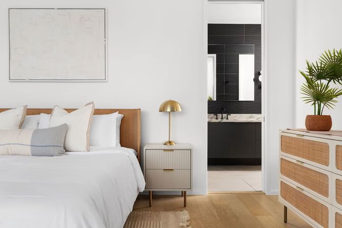 Bright minimalist bedroom with white bedding, rattan headboard, cozy pillows, bedside table with brass lamp, woven-drawer dresser and potted plant, doorway revealing a sleek dark-tiled en-suite bathroom.