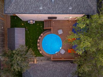 Aerial view of a backyard wooden deck with a round blue above-ground pool, striped lounge cushions, lit string lights, patio umbrella, surrounding trees and storage shed.