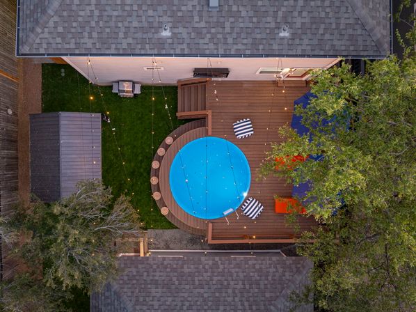 Aerial view of a backyard wooden deck with a round blue above-ground pool, striped lounge cushions, lit string lights, patio umbrella, surrounding trees and storage shed.