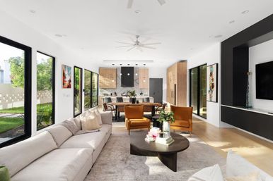 Sunlit modern open-plan living room and kitchen with large black-framed windows, neutral L-shaped sectional, two mustard accent chairs, round dark coffee table with books and plant, wooden dining table and island, light wood floors and white walls.