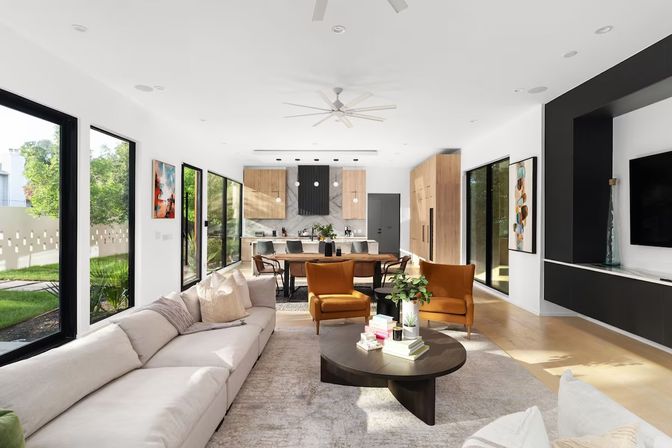 Sunlit modern open-plan living room and kitchen with large black-framed windows, neutral L-shaped sectional, two mustard accent chairs, round dark coffee table with books and plant, wooden dining table and island, light wood floors and white walls.