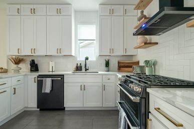 Sunlit modern white kitchen with shaker cabinets and brass hardware, subway tile backsplash, black gas range and dishwasher, marble countertops, floating wood shelves and small potted plants.