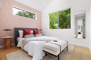Bright modern bedroom with a pink patterned accent wall, gray upholstered bed with red accent pillows and bolsters, white bedding, wooden side tables, plush bench at foot, and a large window framing green trees.
