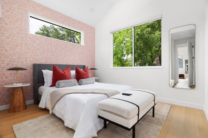 Bright modern bedroom with a pink patterned accent wall, gray upholstered bed with red accent pillows and bolsters, white bedding, wooden side tables, plush bench at foot, and a large window framing green trees.