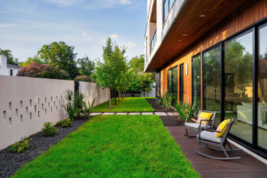 Modern backyard patio with wood-clad exterior and large sliding glass doors, lush green lawn, stepping-stone path, perforated privacy wall, small trees and rock-mulch planting beds, and two outdoor rocking chairs with yellow cushions.