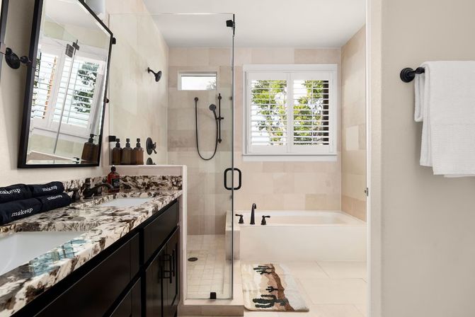 Sunlit modern bathroom with marble-patterned countertop over dark double vanity, frameless glass shower, soaking tub beneath plantation-shutter window, matte-black fixtures, soap dispensers and cactus rug.