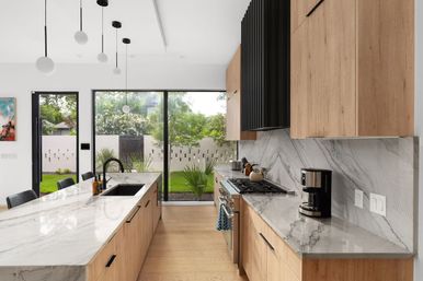 Open-concept modern kitchen with marble waterfall island, light oak cabinets, black pendant lights, gas range and coffee maker, and floor-to-ceiling sliding glass doors showing a lush backyard.
