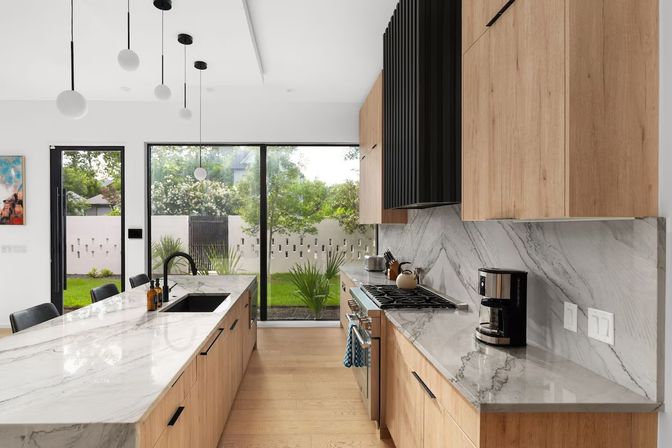 Open-concept modern kitchen with marble waterfall island, light oak cabinets, black pendant lights, gas range and coffee maker, and floor-to-ceiling sliding glass doors showing a lush backyard.