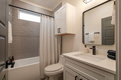 Bright modern residential bathroom with textured gray wave tile shower, white tub and patterned curtain, compact toilet, white vanity and wall cabinet, black fixtures and rectangular mirror — stylish small-home bathroom.
