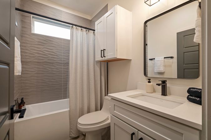 Bright modern residential bathroom with textured gray wave tile shower, white tub and patterned curtain, compact toilet, white vanity and wall cabinet, black fixtures and rectangular mirror — stylish small-home bathroom.