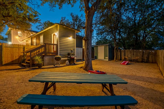 Cozy backyard at dusk with teal wooden picnic table in the foreground, a small lit cottage with a deck and string lights, charcoal grill, cornhole boards, storage shed and trees inside a wooden fence