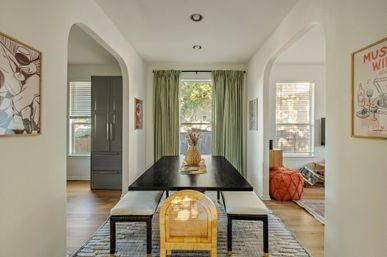 Sunlit modern dining room interior with a black rectangular table, bench seating and a rattan end chair, pineapple centerpiece, floor-length green curtains, arched doorways and hardwood floors.