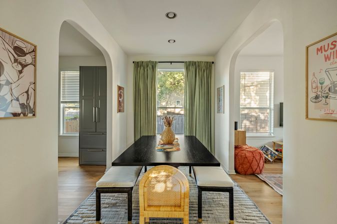 Sunlit modern dining room interior with a black rectangular table, bench seating and a rattan end chair, pineapple centerpiece, floor-length green curtains, arched doorways and hardwood floors.