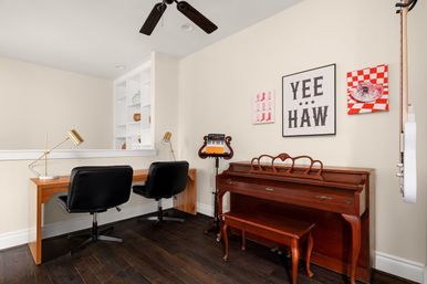 Bright home office and music corner with a wooden upright piano and bench, dual wooden work desk with black swivel chairs and brass lamps, built-in white shelving, dark hardwood floors, ceiling fan, and playful "YEE HAW" wall art.