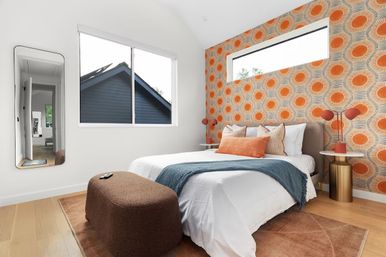 Sunlit modern bedroom with bold orange circular retro wallpaper, white bed dressed with a blue throw and orange accent pillows, brown ottoman, brass side tables with orange lamps and a large window