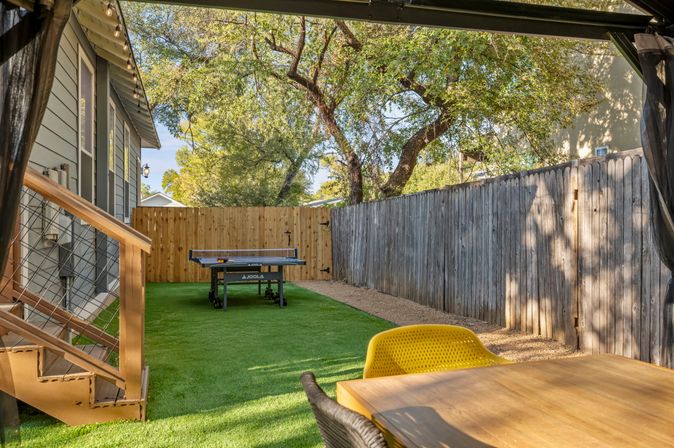 Sunny suburban backyard patio with artificial turf, outdoor ping-pong table, wooden privacy fence, shaded oak trees and a dining table with a yellow chair.