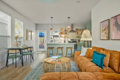 Sunlit open-plan living room and kitchen featuring an orange velvet sectional with teal pillow, round rattan coffee table, green island with three bar stools, subway tile backsplash and pendant lights.
