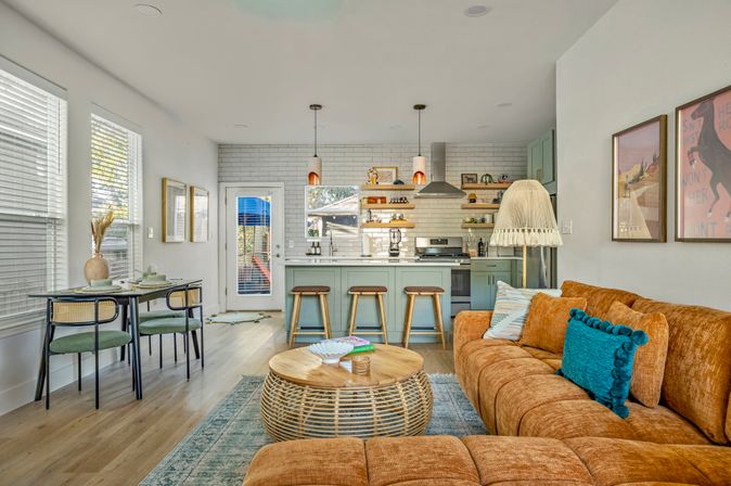 Sunlit open-plan living room and kitchen featuring an orange velvet sectional with teal pillow, round rattan coffee table, green island with three bar stools, subway tile backsplash and pendant lights.